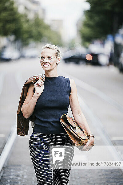 Lächelnde weibliche Fachkraft  die eine Jacke über der Schulter trägt  während sie mit einer Tasche auf der Straße geht