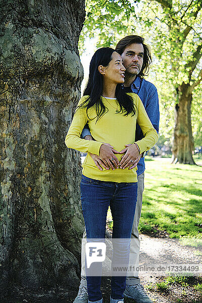 Smiling pregnant woman looking away while talking with husband in front of tree at park