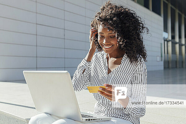 Smiling woman with credit card doing online shopping while sitting outside office building