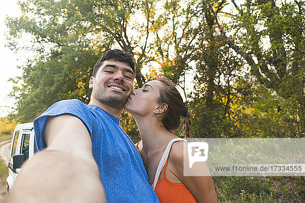 Woman kissing boyfriend while taking selfie at forest