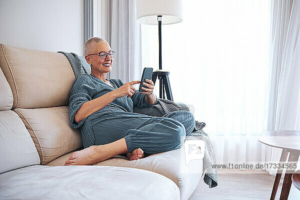 Lächelnde Frau  die ihr Smartphone benutzt  während sie sich auf dem Sofa zu Hause entspannt