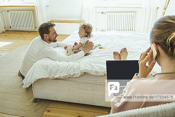 Smiling father and daughter looking at each other while woman talking on mobile phone at bedroom