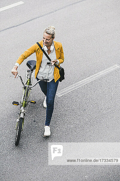 Glückliche Pendlerin  die ihr Mobiltelefon in der Hand hält  während sie mit dem Fahrrad auf der Straße fährt