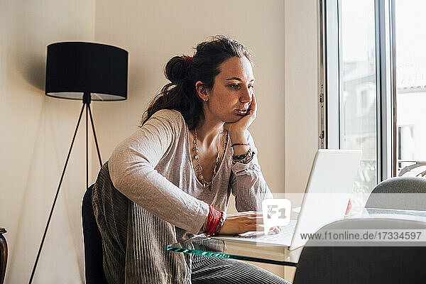 Frau mit Hand am Kinn  die zu Hause sitzend einen Laptop benutzt