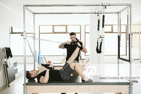 Männlicher Fitnesstrainer stellt den Gurt ein  während ein Mann auf einer Pilates-Maschine im Studio liegt