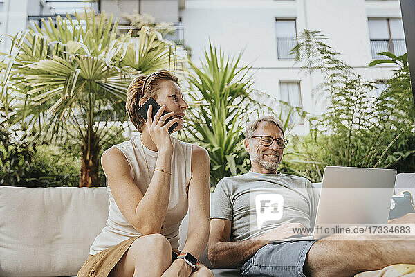 Frau telefoniert mit einem Mann  der auf der Terrasse einen Laptop benutzt
