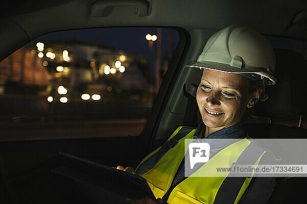Lächelnde weibliche Fachkraft  die nachts im Auto ein digitales Tablet benutzt