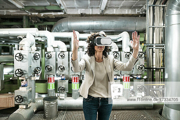 Geschäftsfrau mit erhobener Hand  die ein Virtual-Reality-Headset benutzt  während sie in einem Kraftwerk steht