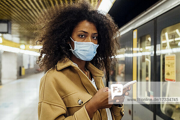Young woman with protective face mask holding mobile phone while standing in subway