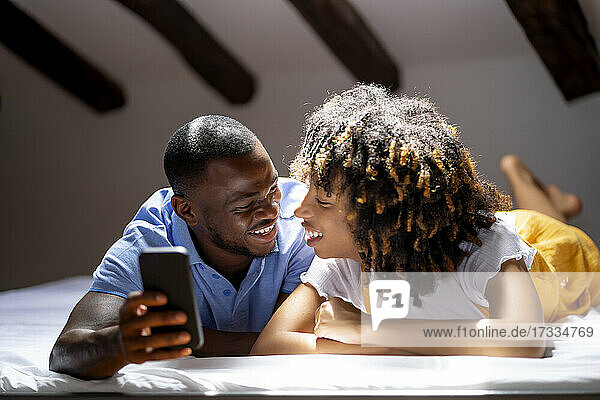 Lächelnder Mann mit Smartphone  der eine Frau ansieht  während er zu Hause auf dem Bett liegt