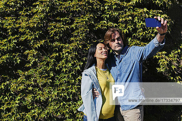 Mann nimmt Selfie mit schwangerer Frau durch Smartphone vor Hecke während sonnigen Tag