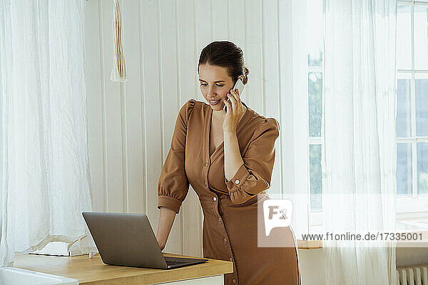 Businesswoman talking on mobile phone while working on laptop at home