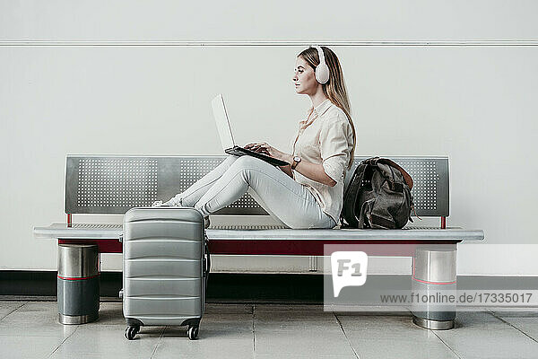Junge Frau  die einen Laptop benutzt und über Kopfhörer Musik hört  sitzt auf einer Bank am Bahnhof