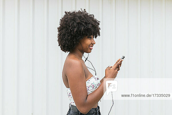 Smiling Afro woman with smart phone listening music through headphones by white wall