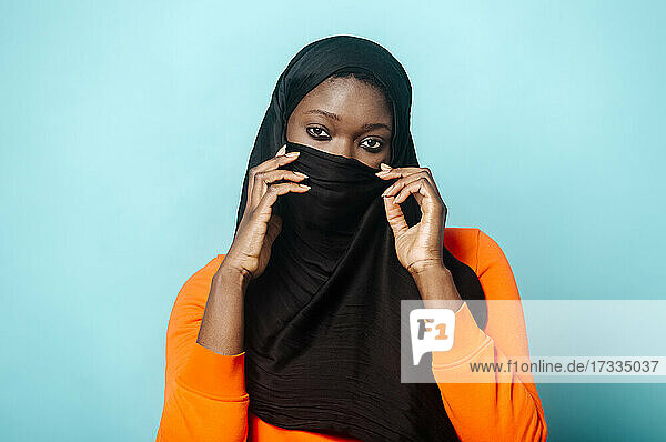 Frau bedeckt Gesicht mit Hidschab vor blauem Hintergrund