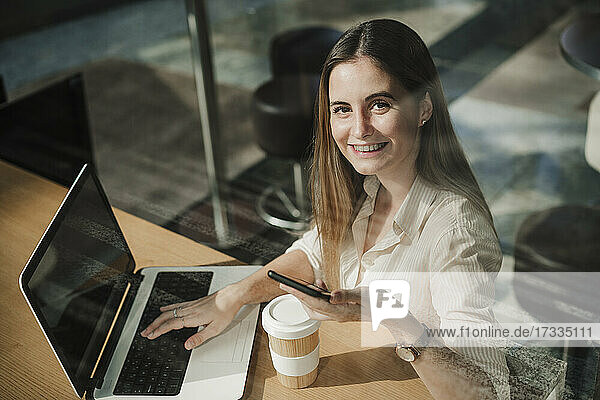 Glückliche junge Freiberuflerin mit Smartphone und Laptop in einem Café