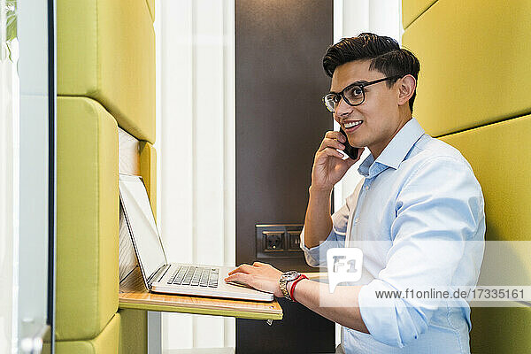 Lächelnder Geschäftsmann mit Laptop  der in einer schalldichten Kabine mit einem Smartphone spricht