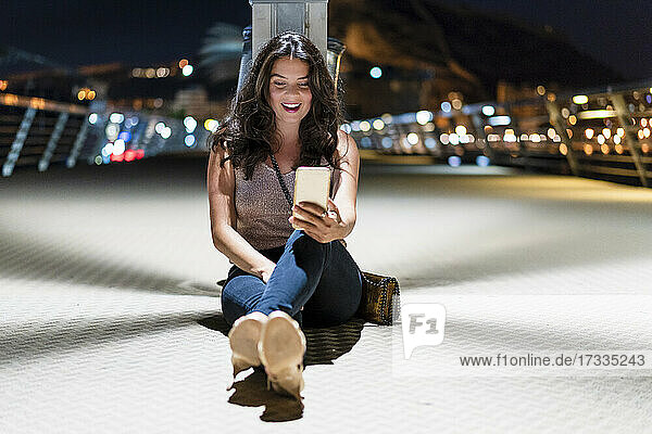 Lächelnde Frau  die ein Mobiltelefon benutzt  während sie nachts auf dem Pier sitzt