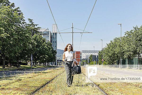 Geschäftsfrau mit Tasche  die an einem sonnigen Tag auf einem Bahngleis spazieren geht