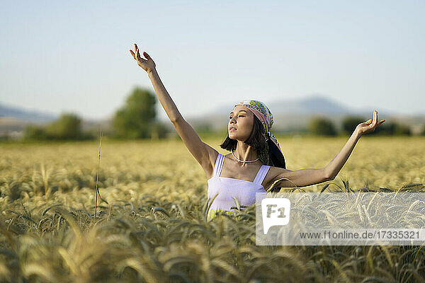 Schöne junge Frau mit Kopftuch steht mit erhobenen Armen in einem Weizenfeld