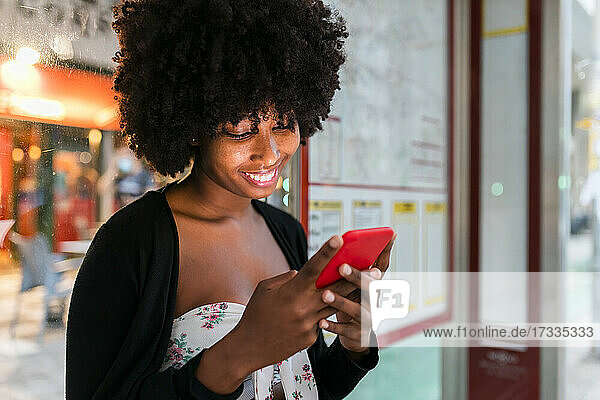 Smiling Afro woman texting message through mobile phone at bus stop
