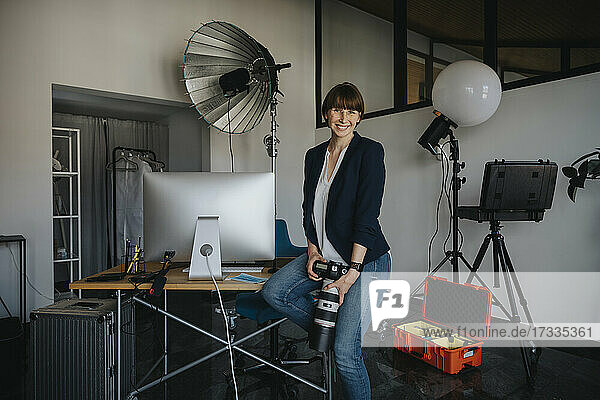 Glückliche weibliche Fachkraft  die eine Kamera hält  während sie auf einem Schreibtisch im Studio sitzt