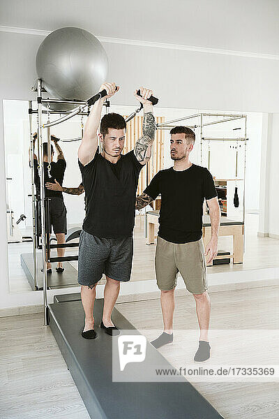 Male fitness instructor standing by man exercising at pilates studio