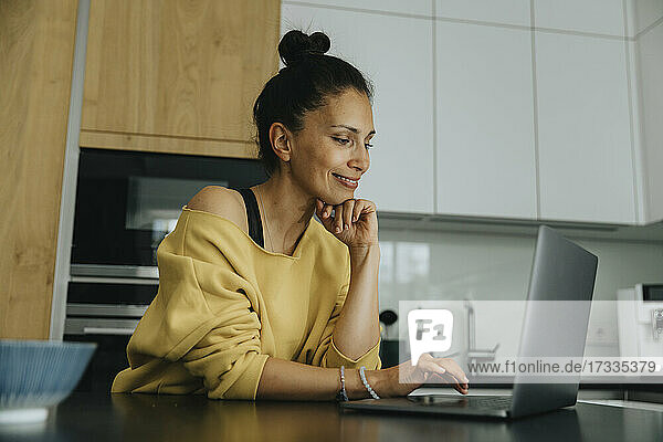 Lächelnde Frau mit Hand am Kinn  die einen Laptop an einer Kücheninsel benutzt