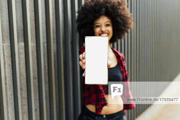 Woman holding mobile phone with blank screen while standing by wall