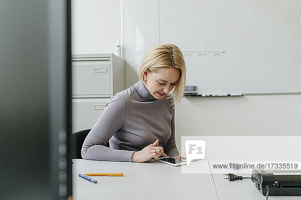 Blonde Geschäftsfrau  die ein Smartphone benutzt  sitzt am Schreibtisch im Büro