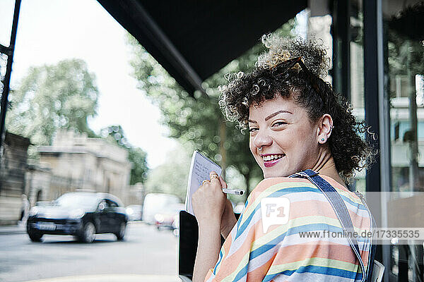 Lächelnde Frau schaut über die Schulter  während sie in einem Straßencafé in einen Notizblock schreibt