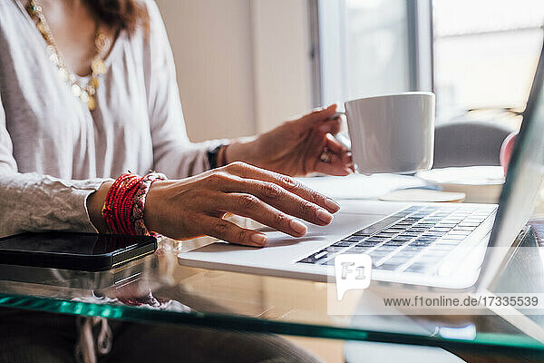 Frau hält Kaffeetasse und benutzt Laptop zu Hause