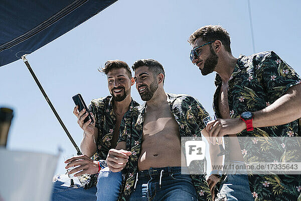 Smiling man sharing mobile phone with male friends in yacht on sunny day during vacation