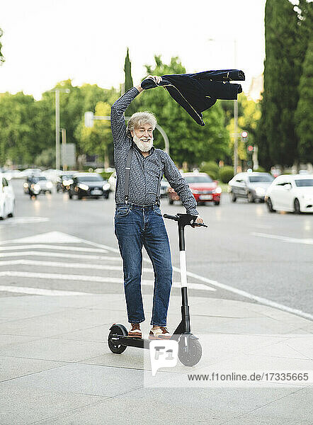 Happy mature man swinging jacket while riding scooter on street