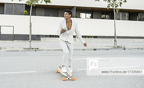 Glücklicher weiblicher Pendler  der sein Smartphone hält  während er auf der Straße Skateboard fährt