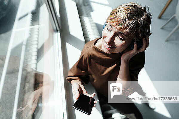 Lächelnde Geschäftsfrau mit Kopfhörern und Mobiltelefon am Bürofenster stehend