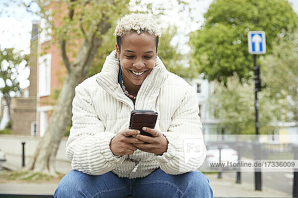 Glückliche erwachsene Frau in weißer Jacke  die ein Smartphone benutzt und im Park sitzt