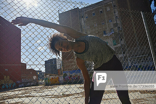Sportswoman doing stretching exercise in front of chainlink fence on sunny day