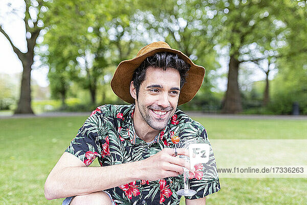Lächelnder Mann mit Hut  der ein Getränk hält  während er im Park sitzt