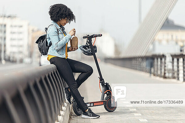Afro-Frau mit Elektroroller isst Essen  während sie sich auf ein Geländer stützt