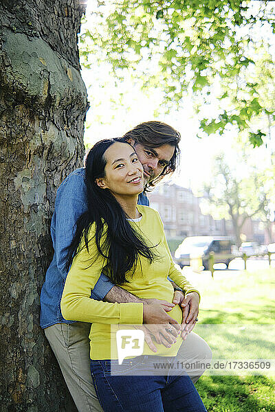 Schwangere Frau lächelt  während ihr Mann sie von hinten im öffentlichen Park umarmt
