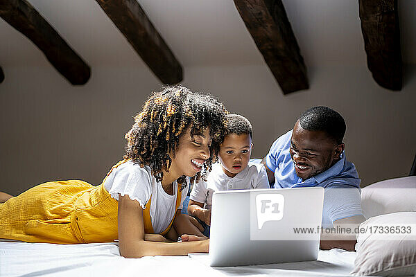 Lächelnde Familie  die einen Laptop mit ihrem Sohn benutzt  während sie zu Hause auf dem Bett liegt