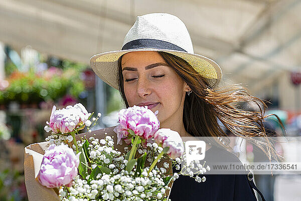 Frau mit Sonnenhut riecht an Pfingstrosen Blumenstrauß in der Nähe von Blumenladen
