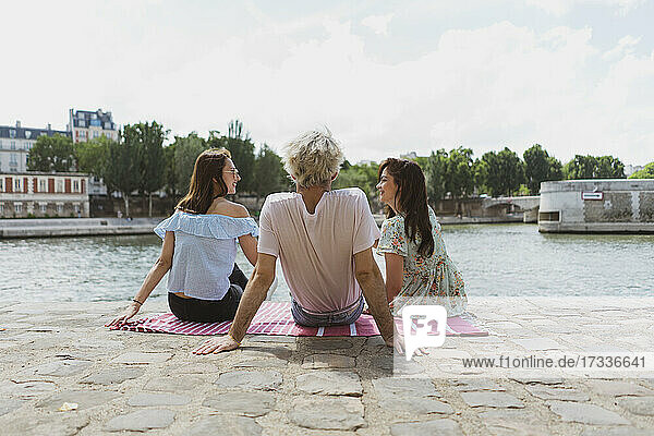 Lächelnde Freundinnen  die sich mit einem Mann unterhalten  während sie auf einer Decke am Fluss Seine sitzen