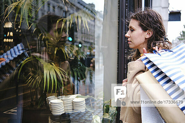 Woman looking through glass window of store while carrying shopping bags