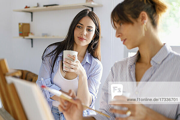 Female artist holding mug looking at colleague painting in studio