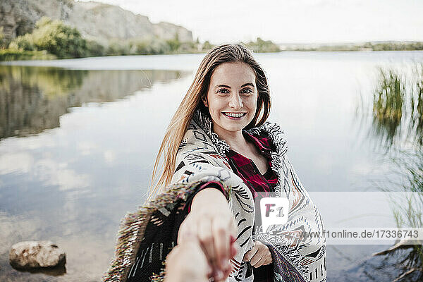 Glückliche junge Frau in eine Decke gewickelt  die die Hand einer Freundin am Seeufer hält