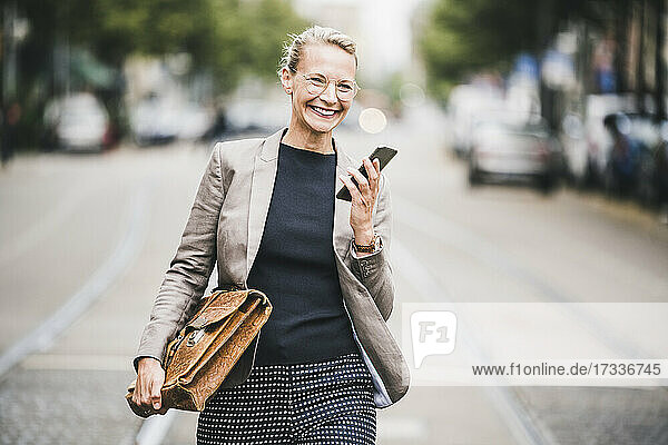 Cheerful businesswoman with smart phone and bag walking on street