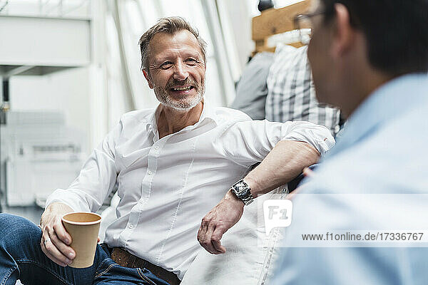 Smiling male professional holding disposable cup while sitting with colleague at office