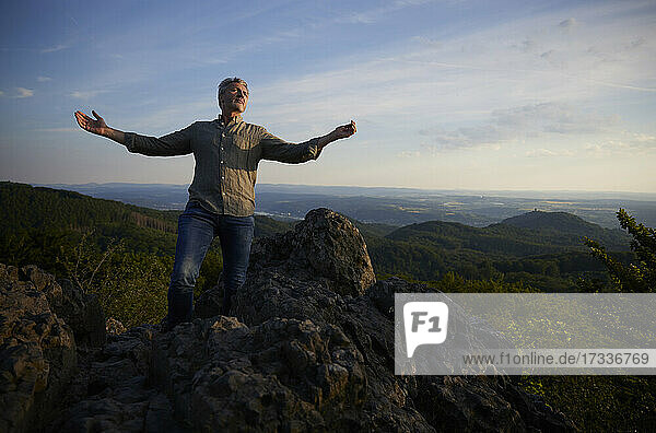 Unbeschwerter Mann mit ausgestreckten Armen auf einem Berg bei Sonnenuntergang stehend